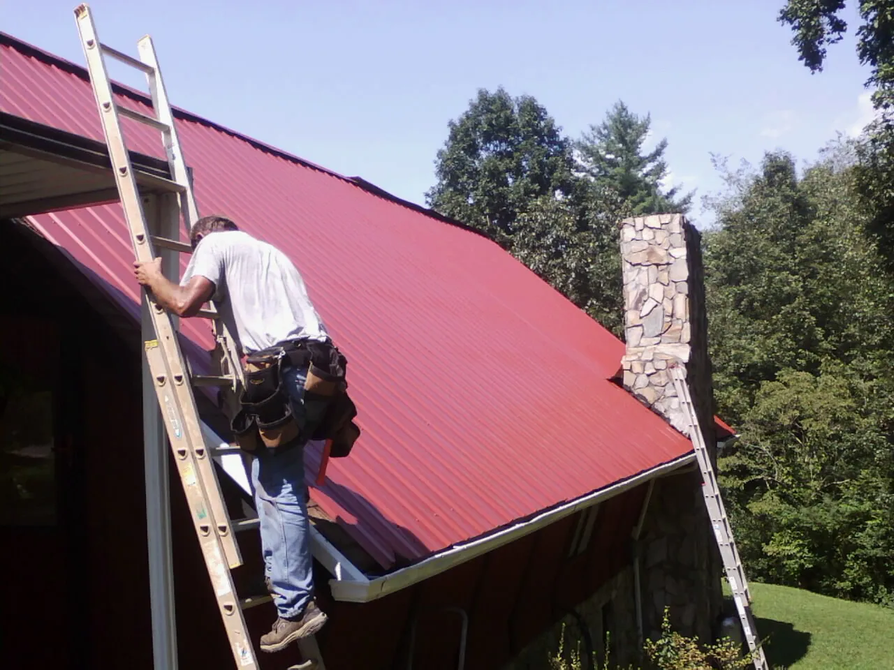 Roofer on a ladder beside a red metal roof and stone chimney — Soffit & Fascia Repair by Roofing Brothers in Eau Claire